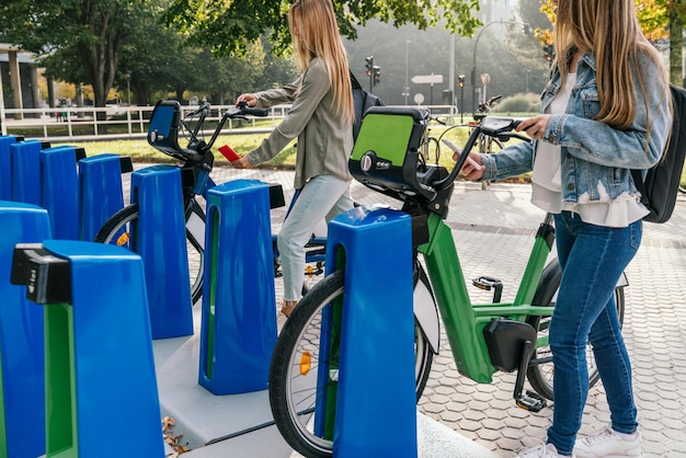 Vista lateral de dos mujeres que utilizan su teléfono inteligente para alquilar dos bicicletas en la estación de alquiler de bicicletas para pasear por la ciudad.