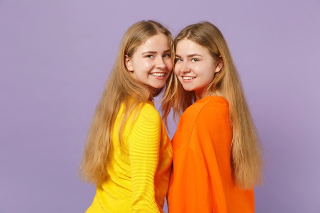 Vista lateral de dos niñas hermanas gemelas rubias jóvenes sonrientes en ropa de colores vivos mirando la cámara aislada en la pared azul violeta pastel. Concepto de estilo de vida familiar de personas.