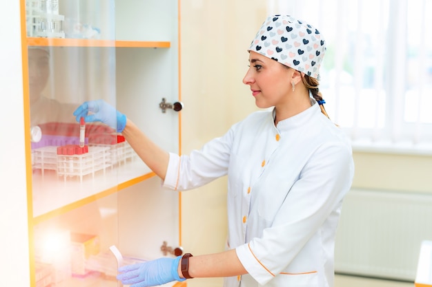 Vista lateral de una enfermera en uniforme médico poniendo tubos de ensayo en el estante. Enfermera profesional vestida con bata blanca, gorro y guantes de látex colocando muestras de sangre en un laboratorio médico.