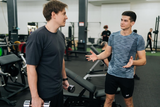 Vista lateral de un entrenador personal masculino ayudando a un joven deportista principiante y discutiendo juntos en el gimnasio