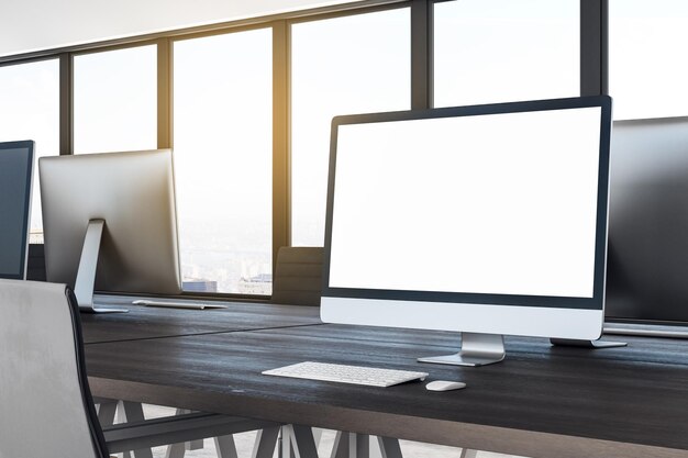 Vista lateral del escritorio creativo de diseño de madera con pantalla de computadora blanca vacía y ventanas brillantes con vista a la ciudad y luz solar en la oficina de coworking Concepto de lugar de trabajo Maqueta de representación 3D