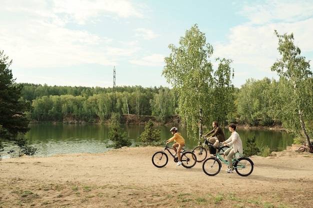 Vista lateral de la familia joven sentada en bicicletas y moviéndose a lo largo de la orilla del río en árboles verdes mientras disfrutan juntos de un descanso activo