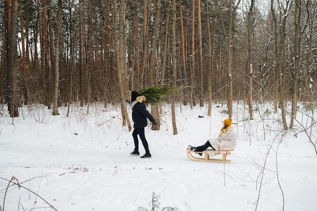 Vista lateral de la feliz pareja. Un hombre con un árbol de Navidad al hombro camina por el bosque de invierno y tira de un trineo con una mujer detrás de él.