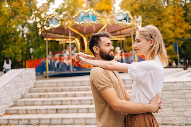 Vista lateral de la feliz pareja joven enamorada de pie abrazándose sonriendo mirándose el uno al otro en el parque de atracciones en el día de verano