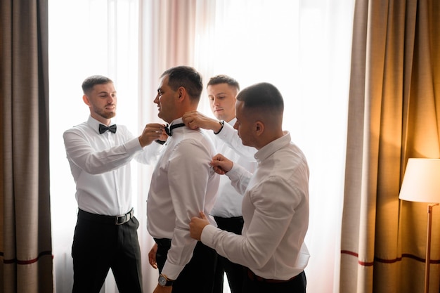 Vista lateral de un grupo de amigos parados rodeados de novios ayudándolo a usar traje y corregir la corbata de moño en su cuello durante la mañana de la boda en apartamentos de lujo