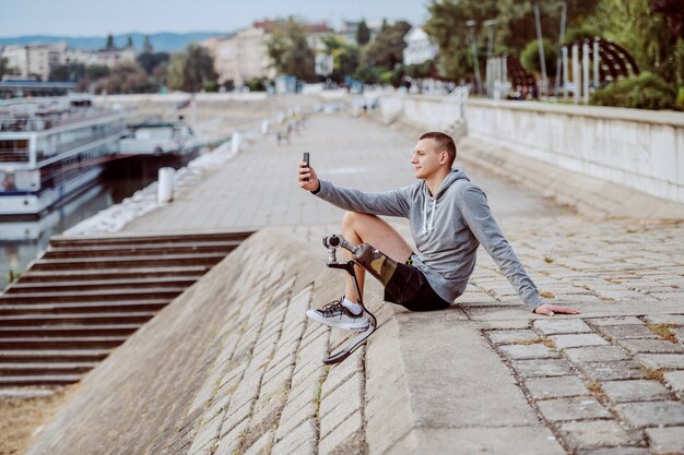 Vista lateral del guapo deportista discapacitado caucásico en ropa deportiva y pierna artificial sentado en el muelle y tomando selfie.