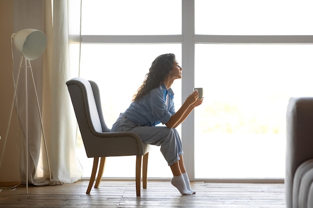 Vista lateral de una hermosa joven bebiendo café caliente sentada en una silla cerca de la ventana en casa gratis