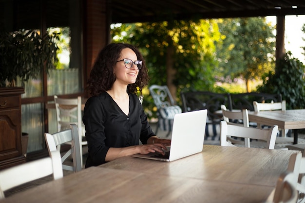 Vista lateral de una hermosa joven con cabello rizado sentada en la mesa escribiendo en la computadora portátil en la terraza