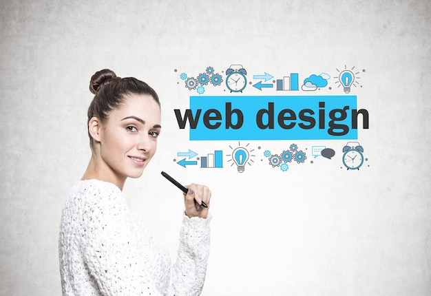 Vista lateral de una hermosa joven morena con un suéter blanco sosteniendo un marcador y sonriendo. Un fondo de pared de hormigón con un boceto de diseño web.