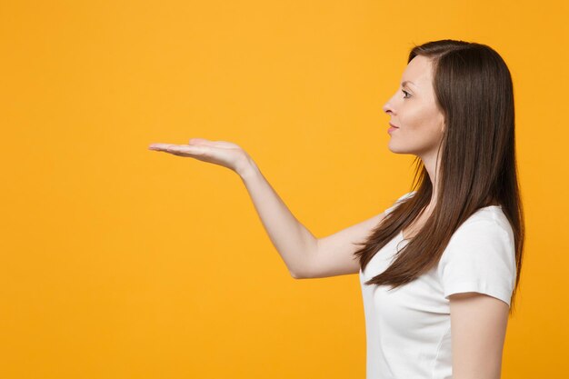 Vista lateral de una hermosa joven con ropa informal blanca que señala la mano a un lado, sostenga algo en las palmas aislado en un fondo de pared naranja amarillo brillante. Concepto de estilo de vida de las personas. Simulacros de espacio de copia.