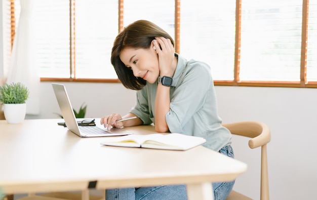 Vista lateral de una hermosa mujer independiente asiática que usa auriculares y sonríe mientras mira una laptop sentada en el escritorio en casaTrabajo remotoElearning Nuevo concepto normal