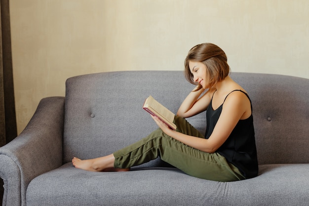 Vista lateral de hermosa mujer con pelo corto, sentado en el sofá gris y leyendo el libro, en casa.