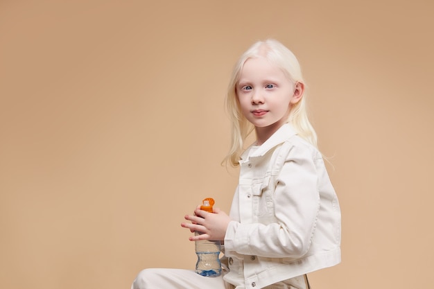 Vista lateral del hermoso niño albino tranquilo y tímido sentado con una botella de agua aislada
