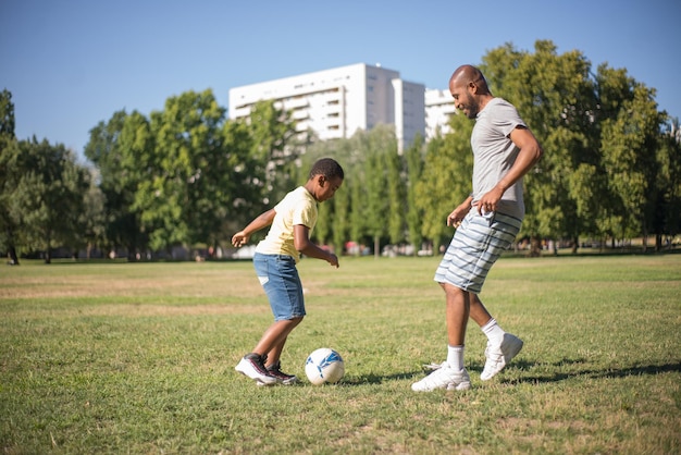 Vista lateral del hombre africano y niño feliz jugando a la pelota en el campo. Padre cuidadoso y su pequeño niño pateando la pelota riendo y divirtiéndose juntos en verano. Actividad deportiva y concepto de estilo de vida saludable.