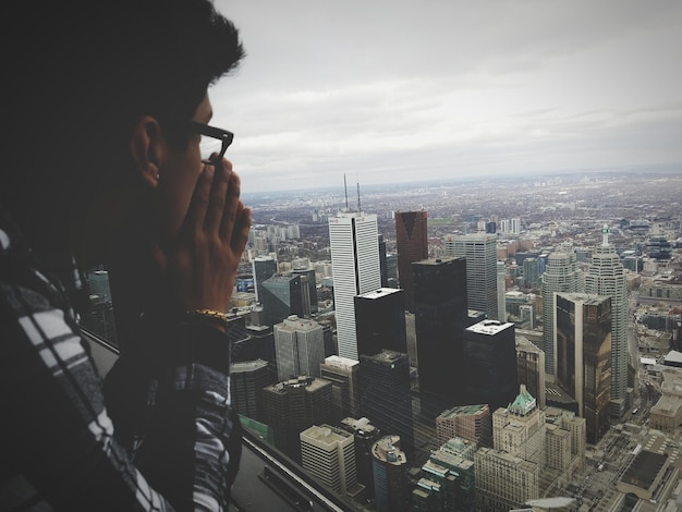 Foto vista lateral de un hombre apoyado en el alféizar de la ventana mientras mira la ciudad contra el cielo nublado