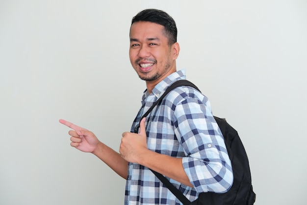 Vista lateral del hombre asiático con mochila sonriendo mientras apunta hacia adelante