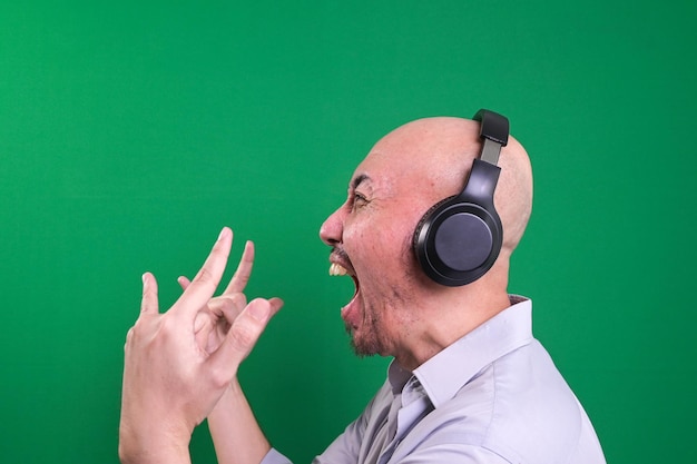 Vista lateral de un hombre calvo asiático escuchando música con auriculares mientras hace un gesto de mano de roca