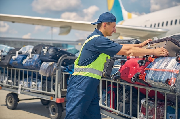 Vista lateral de un hombre caucásico joven serio en uniforme cargando maletas desde el avión en los carros