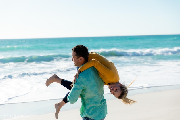 Vista lateral de un hombre caucásico llevando a su novia caucásica sobre su hombro, caminando por la playa con el cielo azul y el mar en el fondo, sonriendo