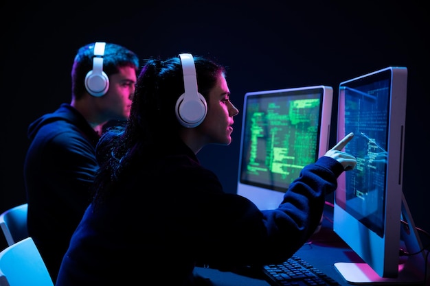 Vista lateral de un hombre caucásico y una mujer trabajando juntos hasta tarde en una oficina por la noche, usando computadoras, programando apuntando y mirando pantallas de computadora, usando auriculares en una habitación oscuramente iluminada