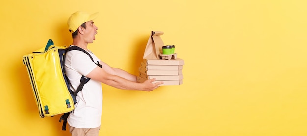 Vista lateral del hombre conmocionado con gorra amarilla y mensajero de camiseta sosteniendo pizza y tazas de papel de café entregando el pedido al cliente posando aislado en fondo amarillo