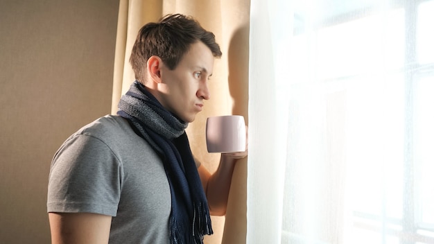 Vista lateral del hombre enfermo en bufanda sosteniendo una taza de bebida aromática caliente mientras está de pie cerca de la ventana en casa