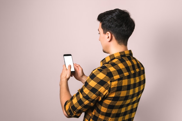 Vista lateral del hombre guapo en camisa amarilla mensajes de texto relajados y chateando con teléfono inteligente contra el fondo neutro