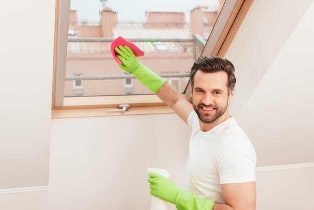 Vista lateral de un hombre guapo sonriendo a la cámara y limpiando la ventana en casa