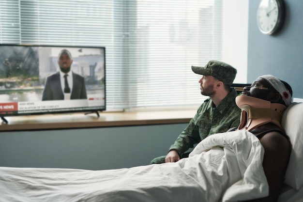 Vista lateral del hombre herido y su amigo en uniforme militar viendo la televisión