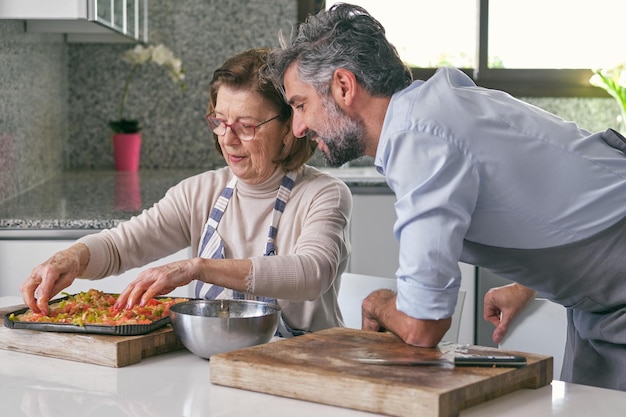 Foto vista lateral de un hombre hispano barbudo maduro con cabello gris y una mujer mayor con delantales preparando deliciosa comida italiana en casa