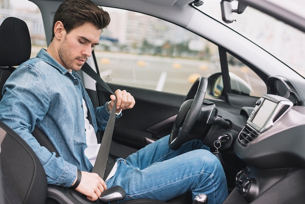 Vista lateral de un hombre joven sentado en el interior del auto que pone el cinturón de seguridad