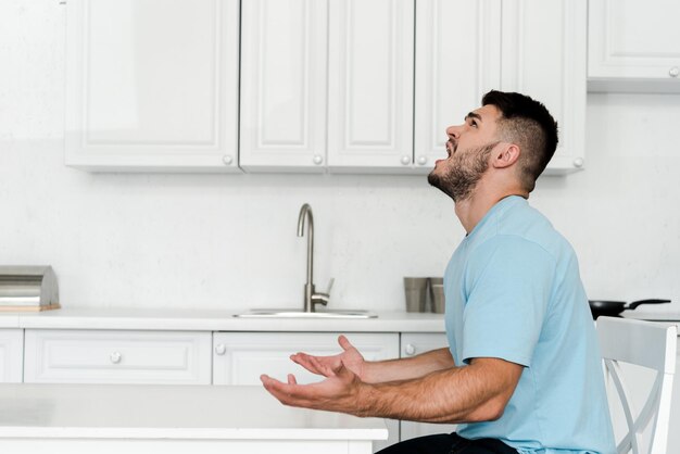 Vista lateral del hombre molesto gritando mientras está sentado cerca de la mesa en la cocina