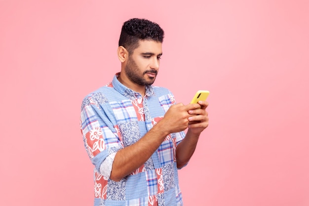 Vista lateral de un hombre moreno serio y concentrado con barba sosteniendo un teléfono celular en las manos chateando o revisando la adicción a los dispositivos de correo electrónico Foto de estudio interior aislada en fondo rosa