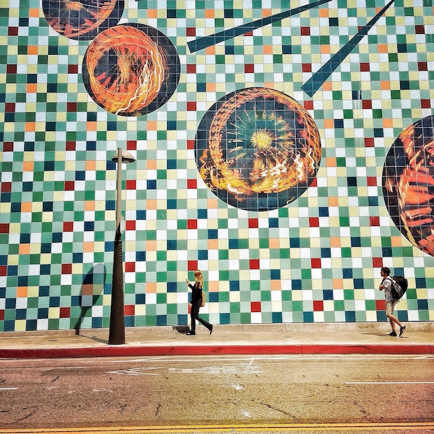 Vista lateral de un hombre y una mujer caminando por la acera contra una pared mural