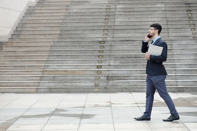 Vista lateral del hombre de negocios caminando al aire libre, llevando una carpeta de documentos y hablando por teléfono con un compañero de trabajo