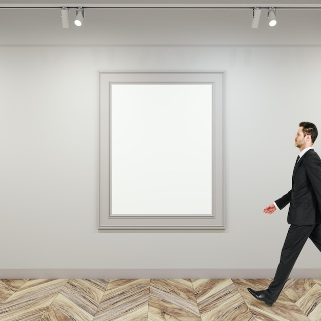 Foto vista lateral del hombre de negocios caminando en el interior de la galería moderna con marco de póster blanco vacío en la pared de hormigón y suelo de parquet de madera