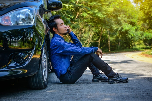 Vista lateral de un hombre de negocios usando el teléfono móvil mientras está sentado contra un coche en la carretera