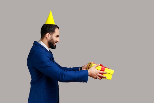 Vista lateral de un hombre positivo con humor festivo parado en un cono de fiesta y dando una caja de regalo, celebrando el cumpleaños de un amigo, usando traje de estilo oficial. Disparo de estudio interior aislado sobre fondo gris.