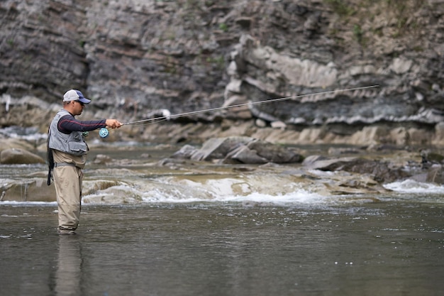 Vista lateral del hombre relajado en ropa impermeable de pie inmóvil en el río mientras pesca. Naturaleza de montaña. Concepto de estilo de vida activo.