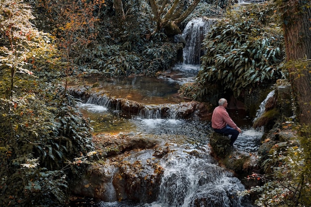 Vista lateral de un hombre sentado en una roca junto a una cascada en el bosque