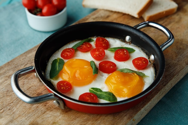 Vista lateral de huevos fritos con tomates decorados con hojas en sartén con rebanadas de pan en tabla de cortar y tomates sobre fondo azul.