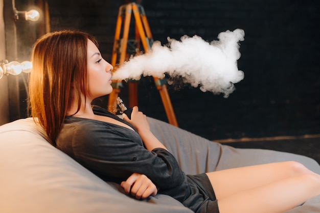 Vista lateral de la impresionante joven usando un cigarrillo electrónico, exhalando una gran nube de humo, pasando el tiempo libre mientras se relaja en el puf de la habitación con un elegante interior.