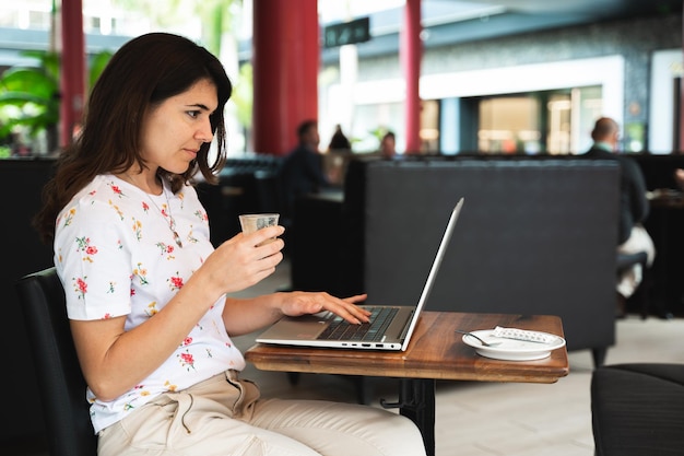 Vista lateral jovem casual sentada à mesa de café usando computador laptop enquanto bebe um café