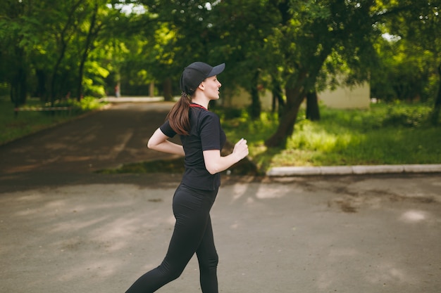 Vista lateral Jovem menina morena atlética de uniforme preto e boné treinando, fazendo exercícios de esporte e correndo, olhando direto no caminho no parque da cidade ao ar livre