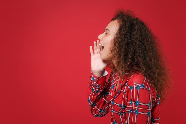 Vista lateral de una joven afroamericana en pijamas descansando en casa aislada en un fondo rojo. Relájese el concepto de estilo de vida de buen humor. Simulacros de espacio de copia. Gritando con gesto de mano cerca de la boca.