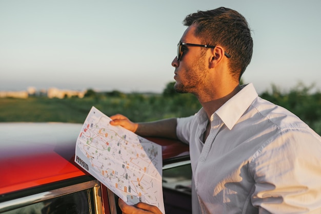 Vista lateral de un joven apuesto que usa un mapa en un viaje por carretera como destino de guía en vacaciones Un turista caucásico mira un mapa mientras está parado al lado del auto en el fondo de la naturaleza al atardecer Viajes