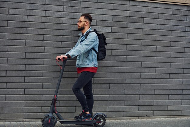 Vista lateral de un joven apuesto con ropa informal que monta una moto eléctrica al aire libre durante el día soleado