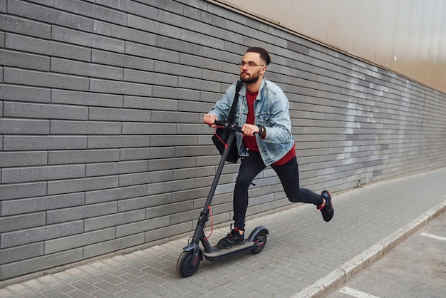 Vista lateral de un joven apuesto con ropa informal que monta una moto eléctrica al aire libre durante el día soleado