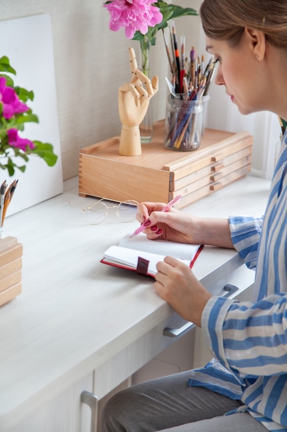 Vista lateral de una joven artista caucásica llena un planificador diario escribe una lista de tareas pendientes en un cuaderno sentado en una mesa de trabajo con cajones con materiales para la creatividad. Concepto de creatividad y afición