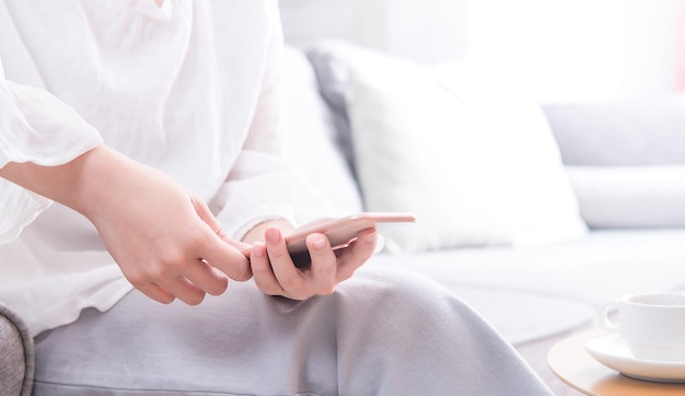 Vista lateral de una joven asiática de blanco sentada en un sofá y usando un teléfono inteligente en un espacio de copia de sala de estar brillante de cerca en blanco para el concepto de diseño